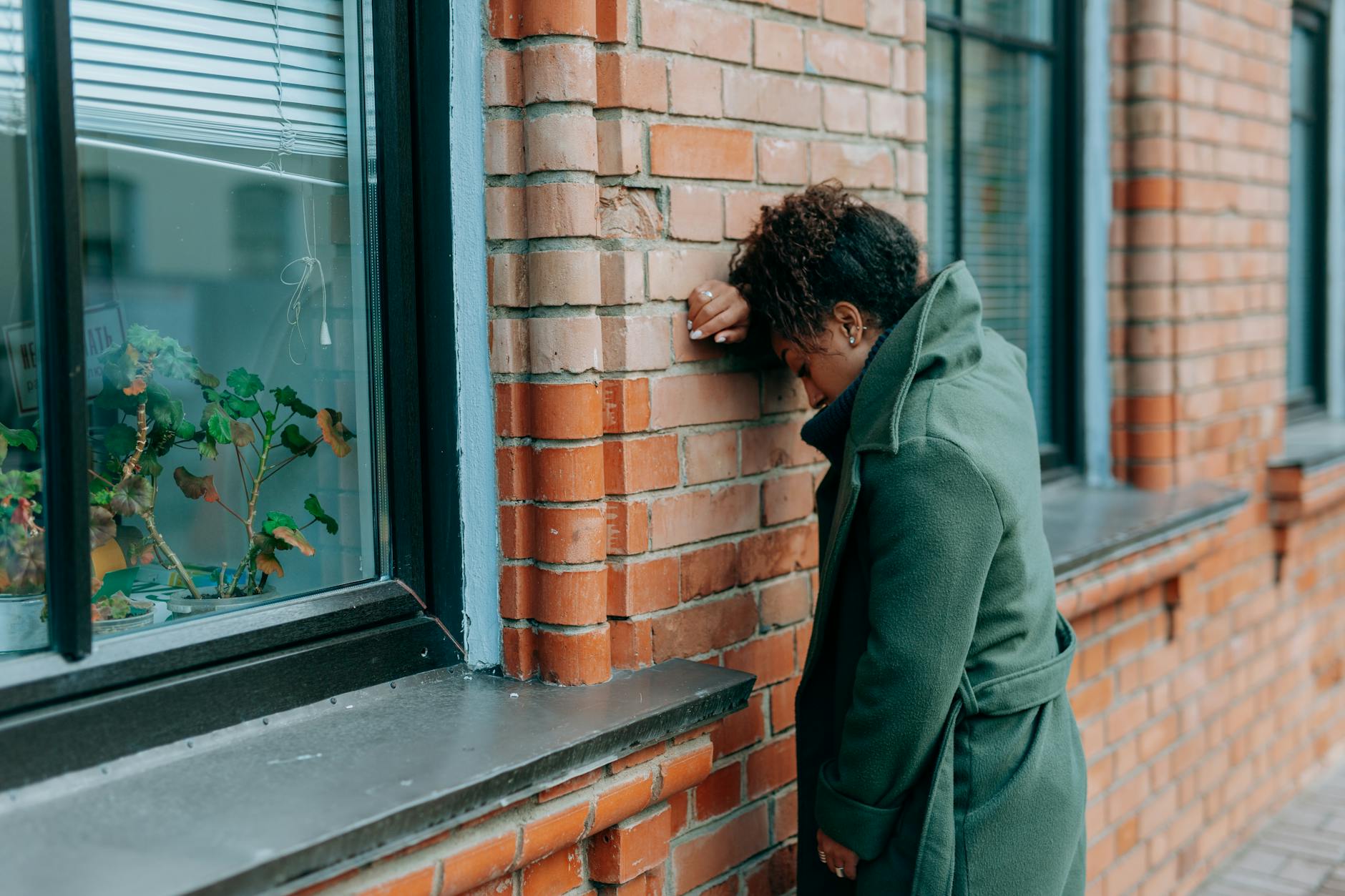 A woman in a green coat leans against a brick wall, reflecting deeply outdoors. - cbt for spring sad