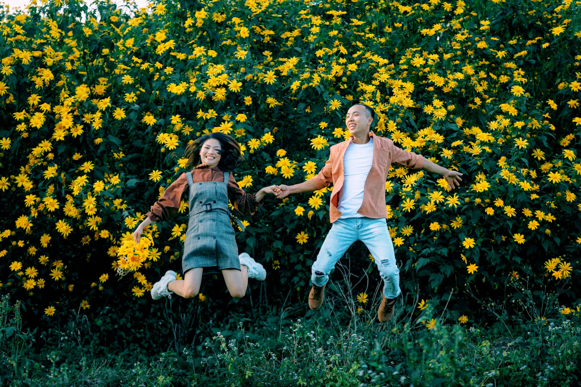 Full body of delighted Asian couple holding hands and jumping above ground near blossoming tree with yellow flowers in nature - cheap spring dates