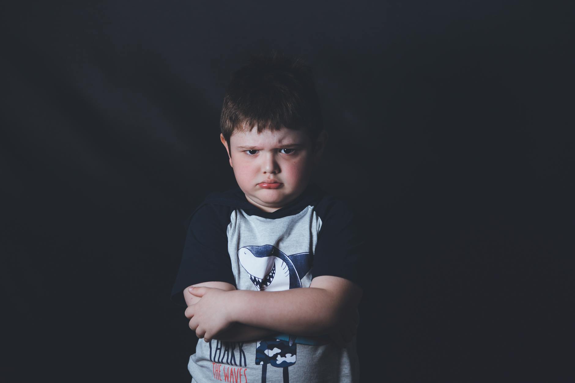 Young boy with arms folded and pouty face against a dark backdrop, showcasing displeasure. - child emotional intelligence