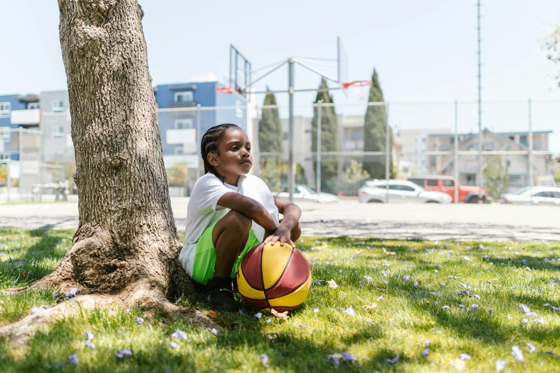 A young child sitting under a tree holding a basketball in a park setting. - child resilience summer