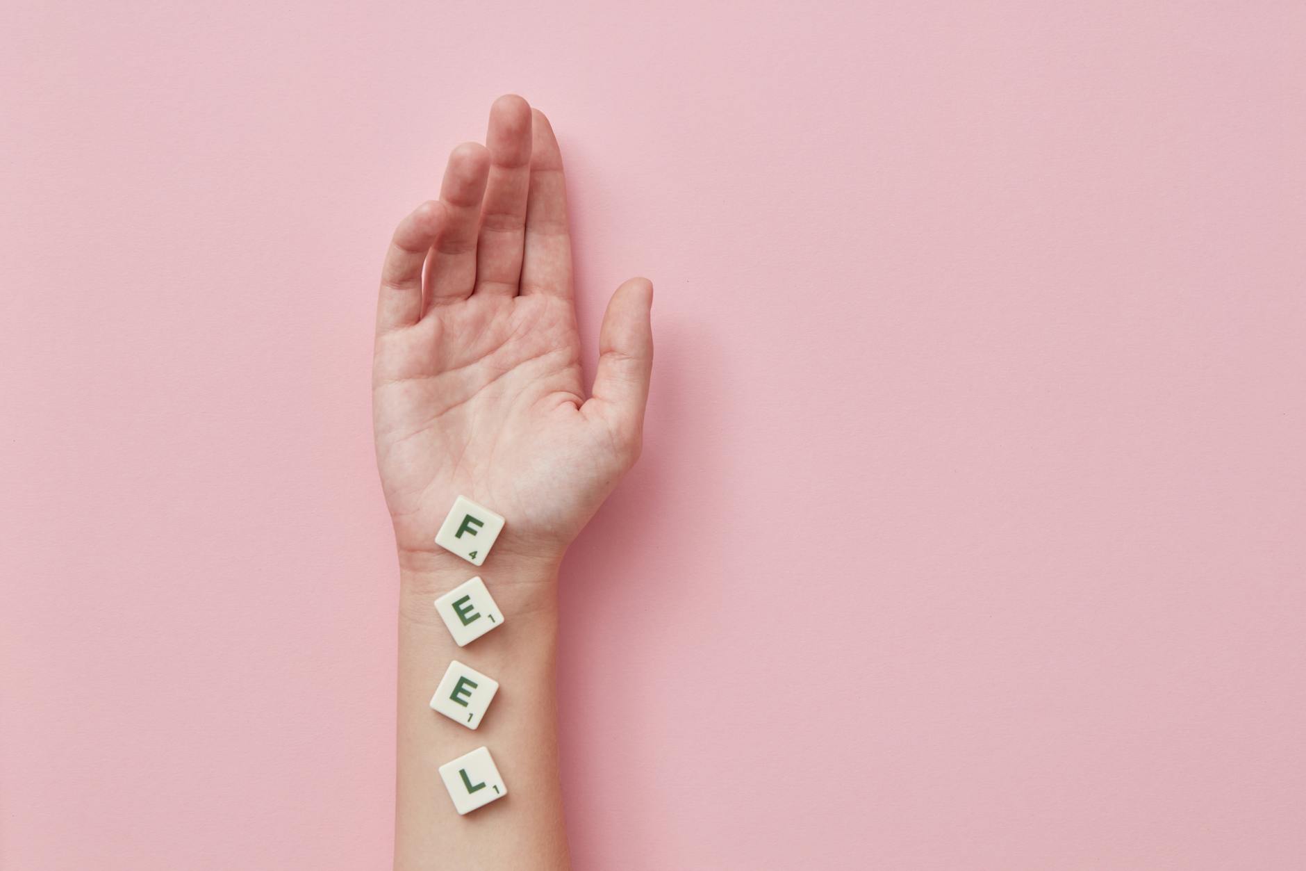 A minimalist photograph of a hand laid on pink background spelling 'FEEL' with Scrabble tiles. - dbt emotional regulation