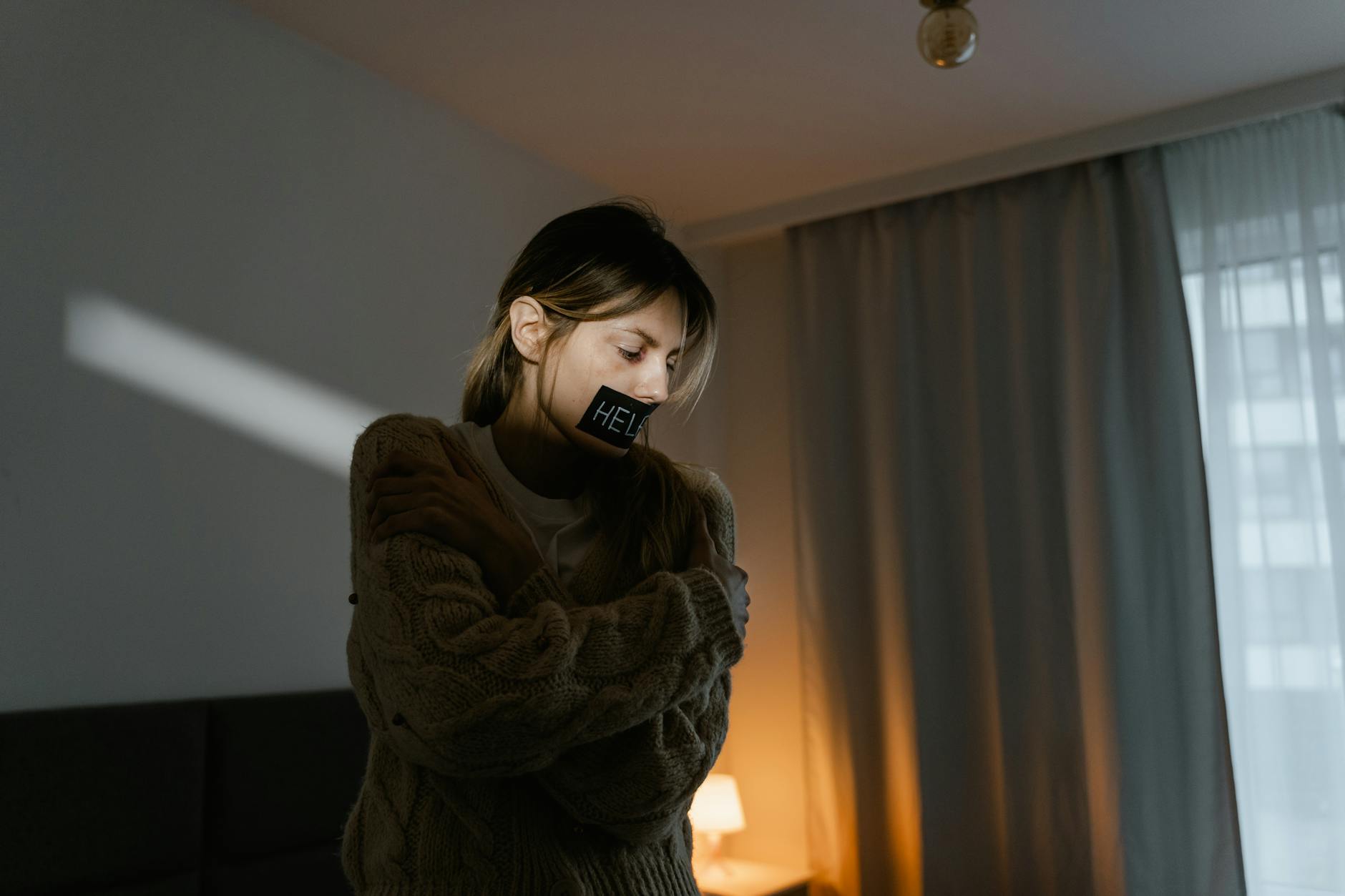 A young woman in a dim room wears a sweater and a help sign, expressing need. - express needs at home