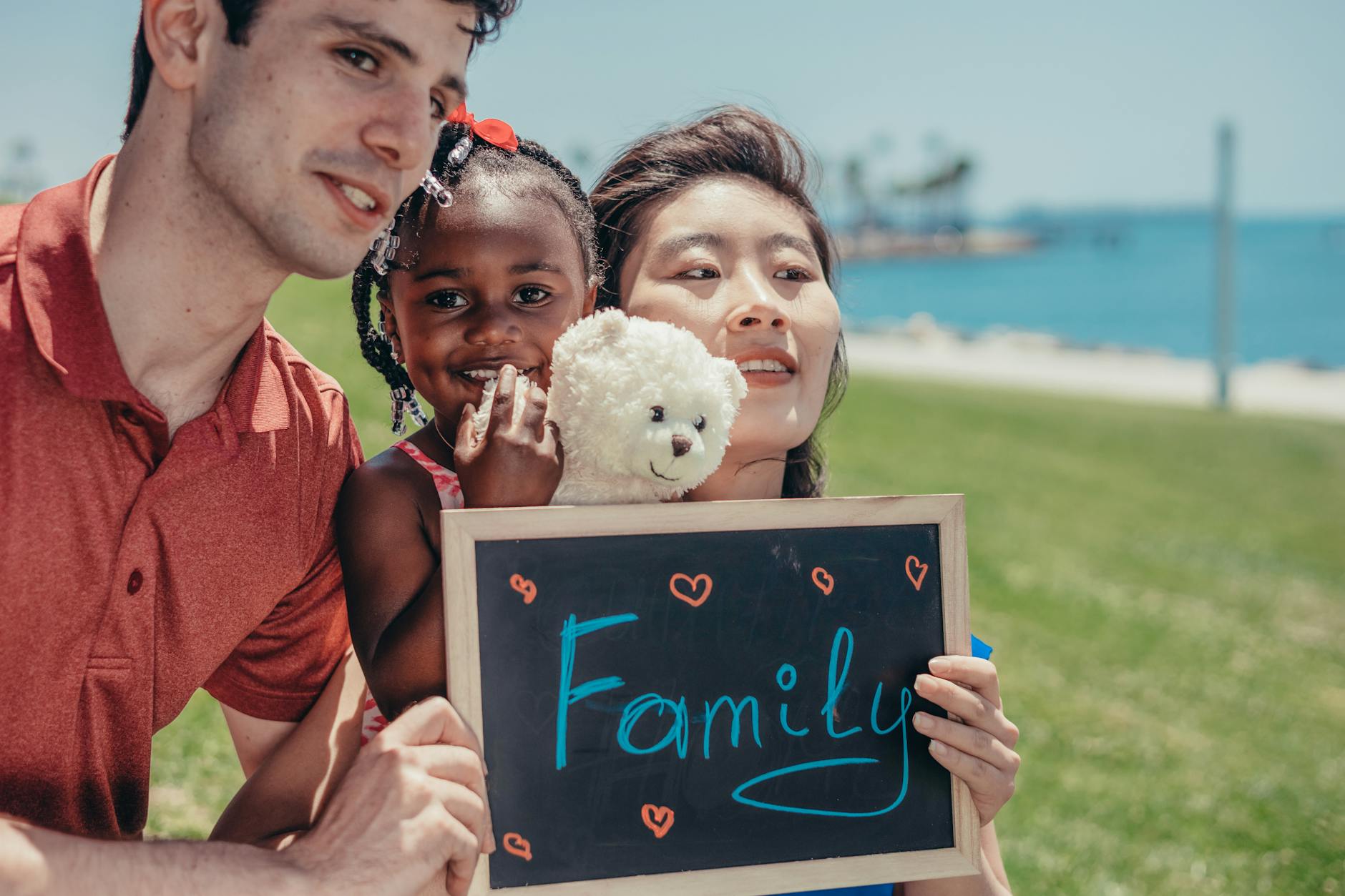Happy mixed family bonding with a teddy bear and a chalkboard that reads 'Family' on a sunny day by the sea. - family summer plans