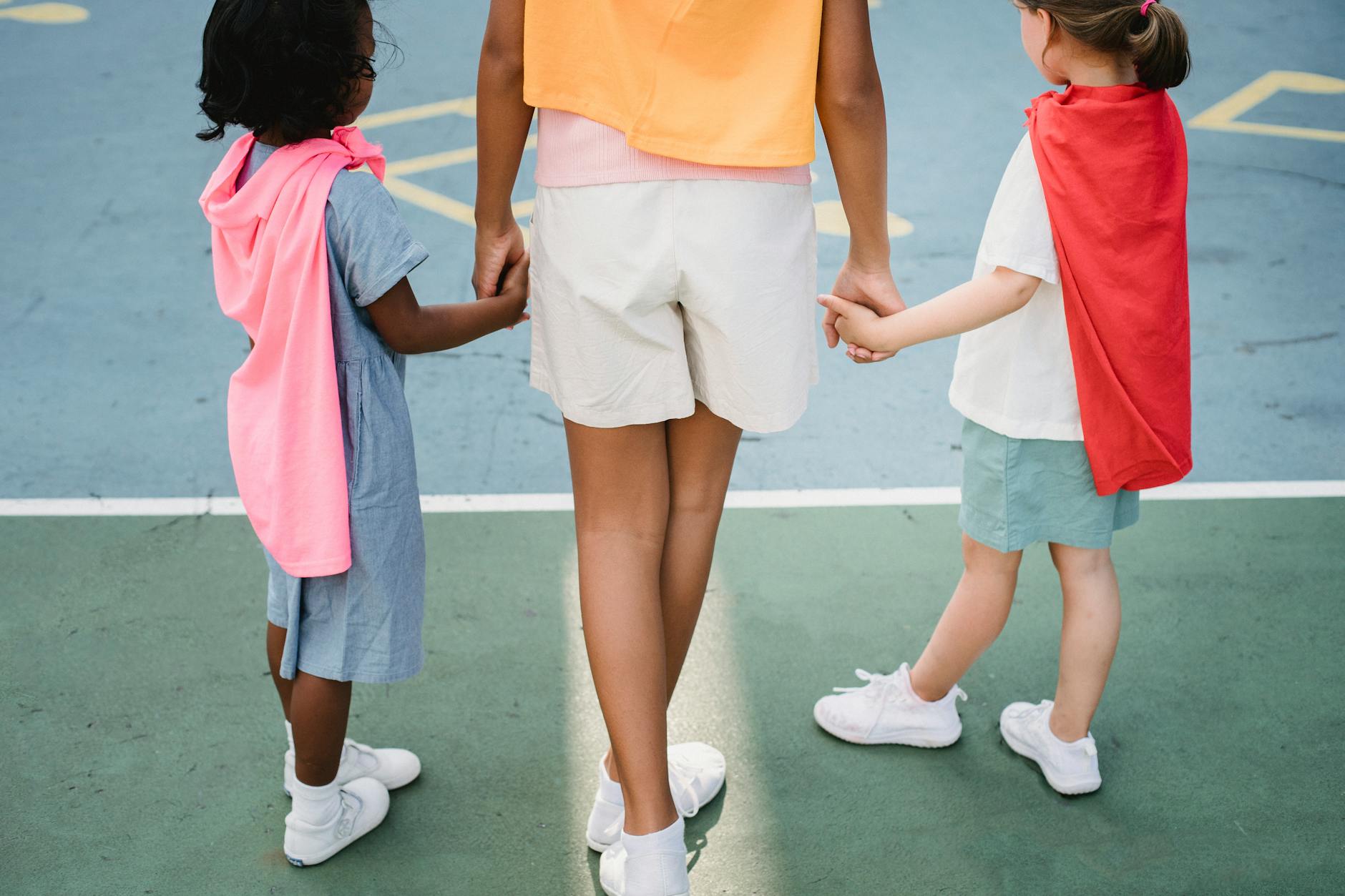 Rear view of children holding hands wearing superhero capes on a playground. - independent outdoor play