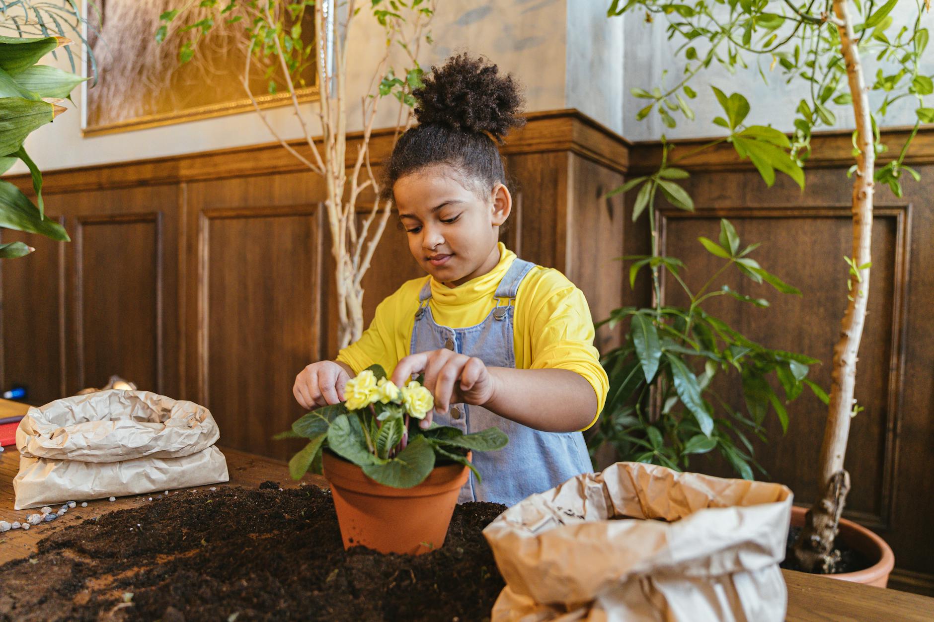 A young girl smiling while potting a plant indoors, surrounded by greenery and soil. - kid resilience spring transition