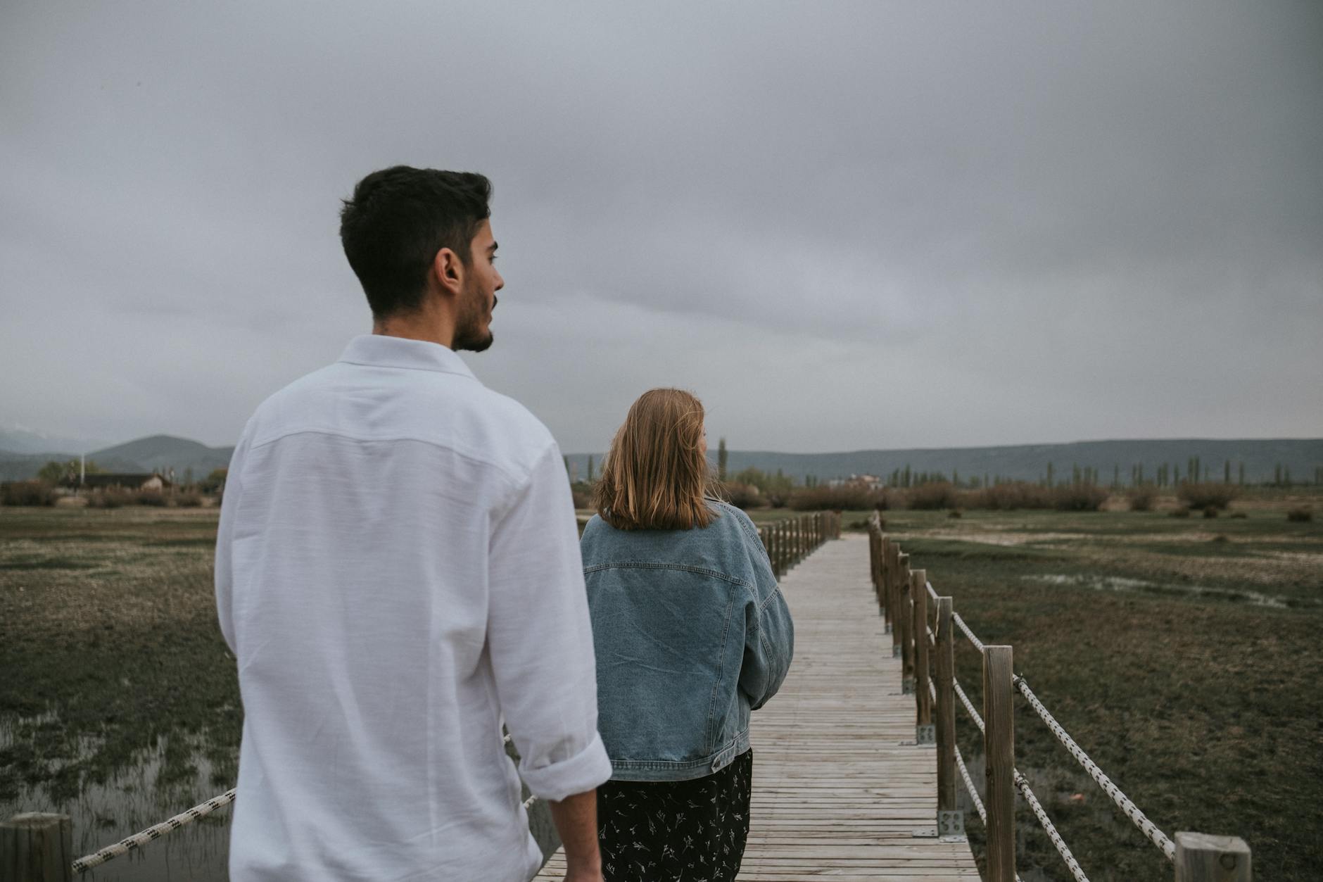 A couple strolls on a wetland footbridge amidst serene nature under overcast skies. - long distance relationship tips