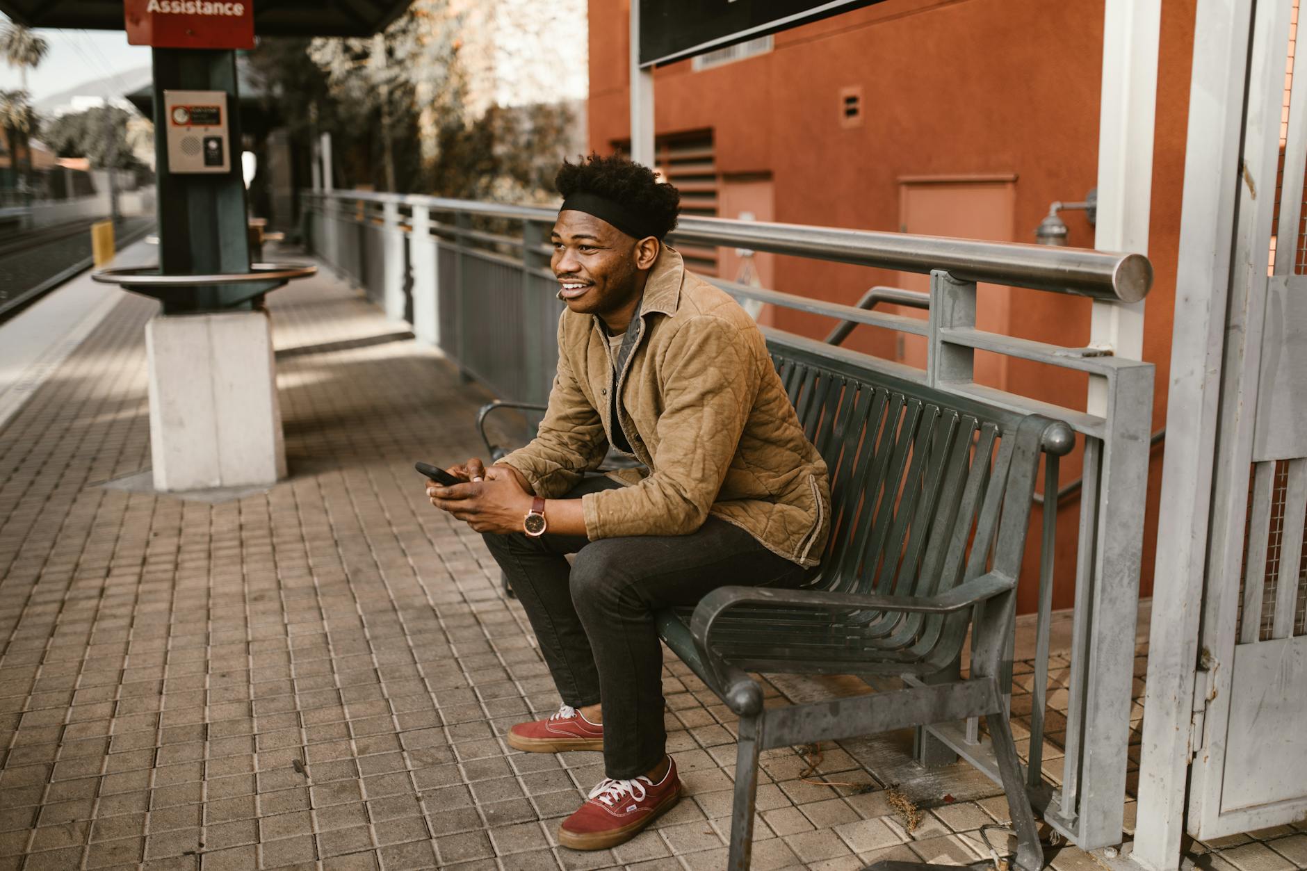 A man sitting on a bench at a city train station while using his phone. - long distance summer