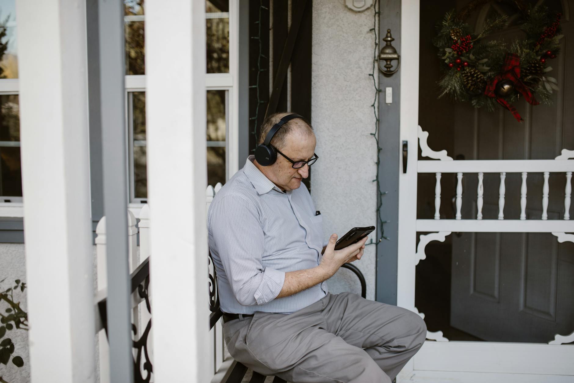 Elderly man using smartphone and headphones on porch. Winter decor suggests festive mood. - long distance summer