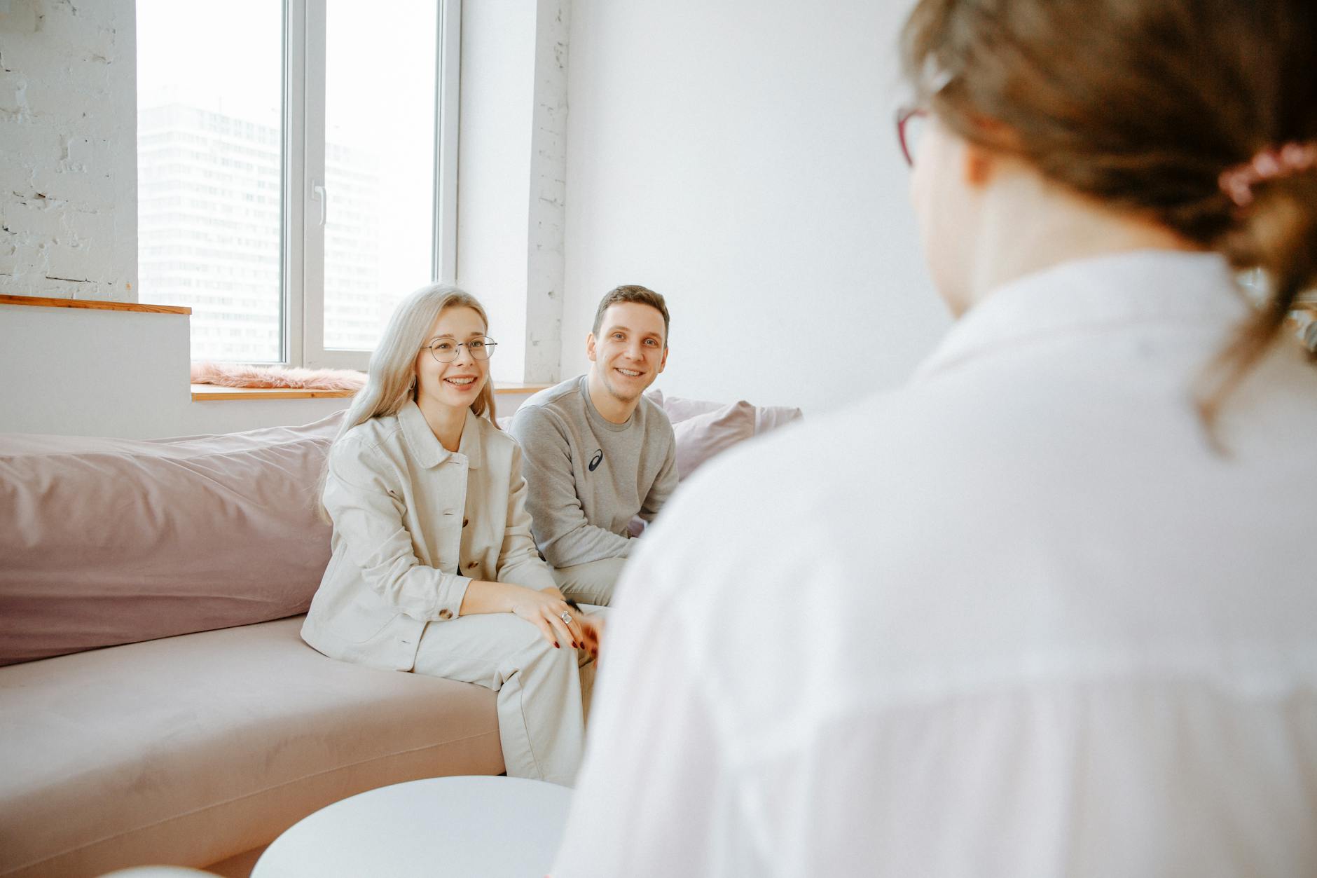 A couple smiles while sitting on a sofa during a counseling session indoors. - marriage counselor questions