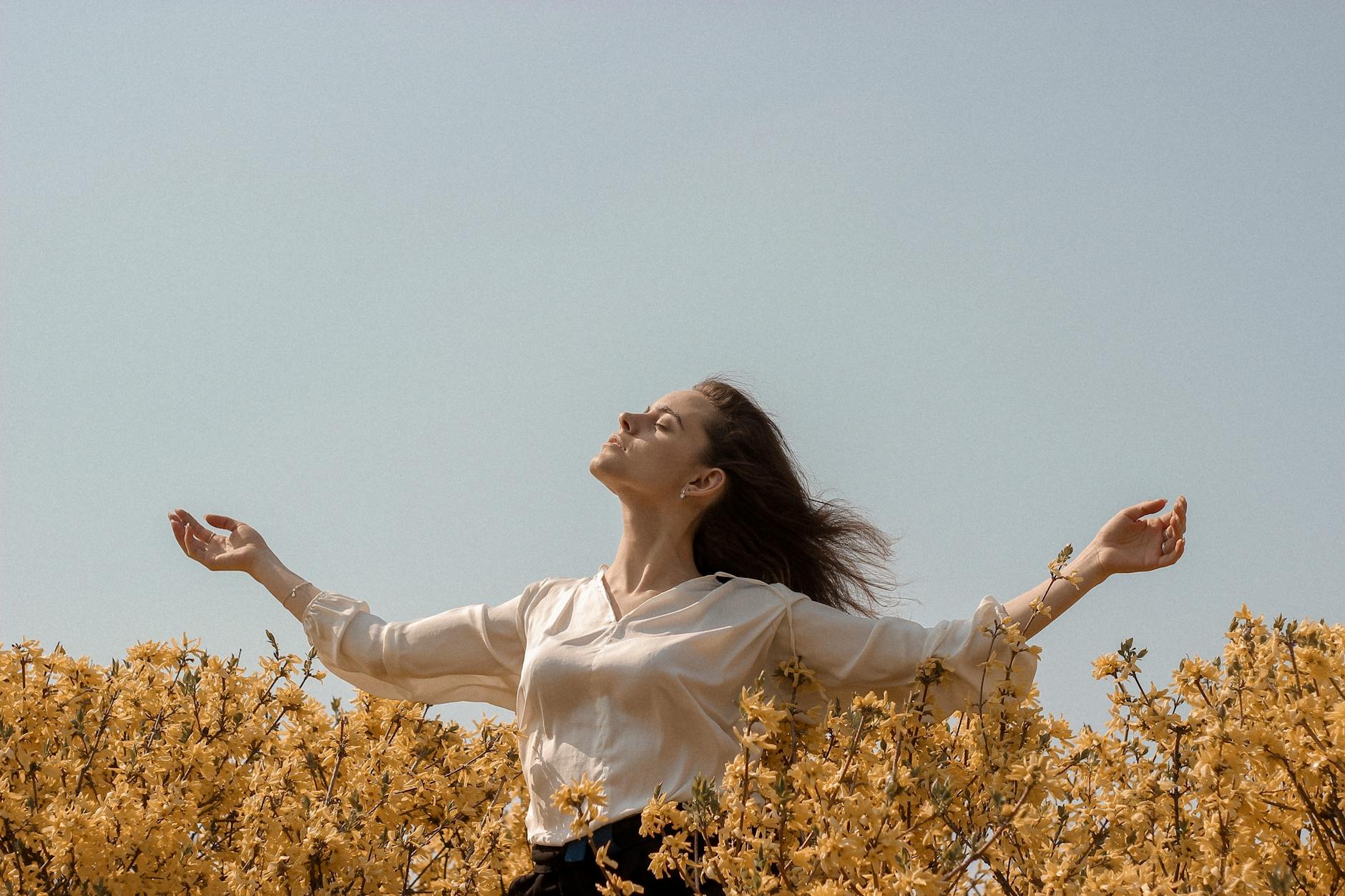 A woman enjoys the serene outdoors with open arms amid vibrant yellow flowers under a clear sky. - mental resilience spring