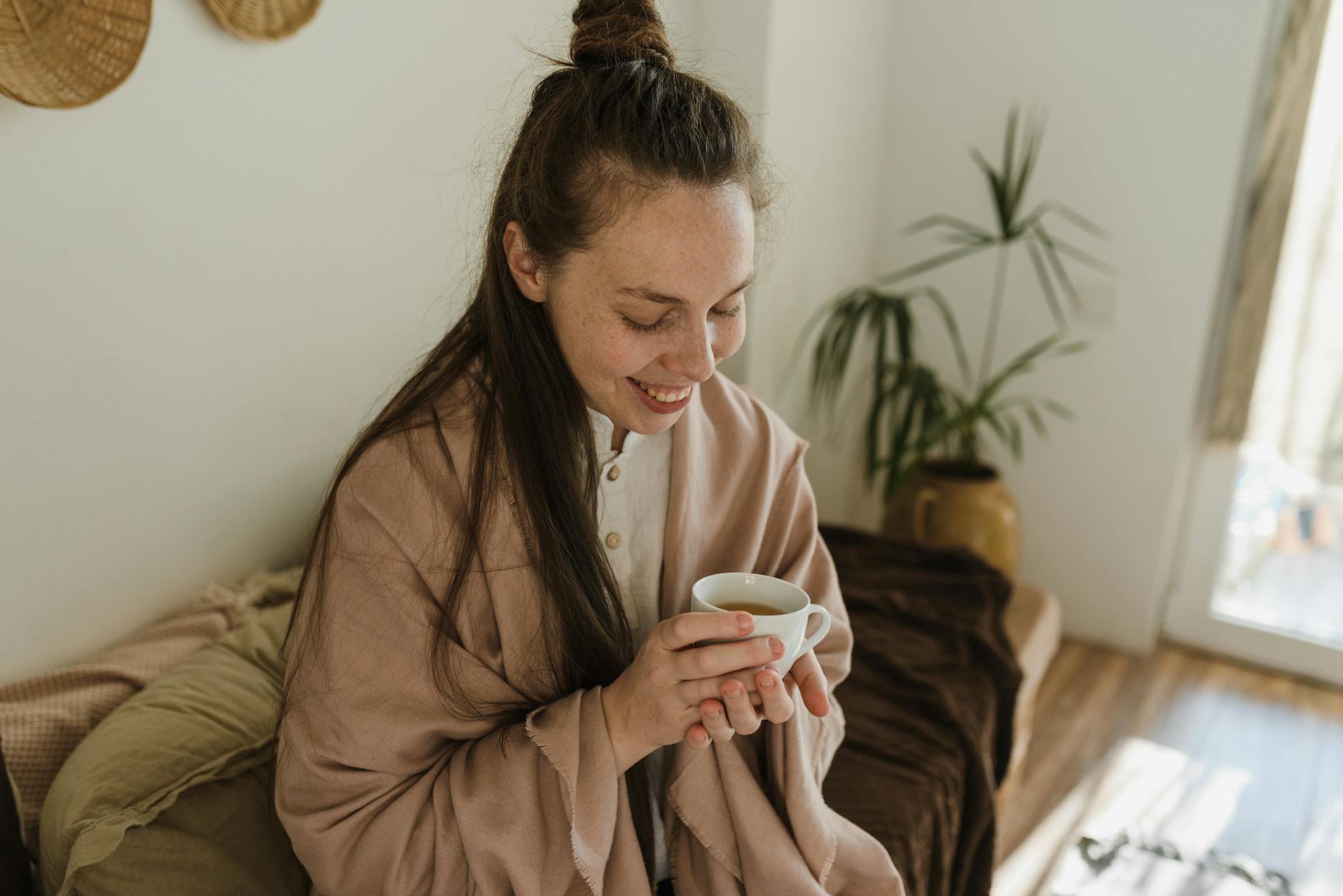 Smiling woman wrapped in a shawl holding a hot cup of coffee indoors. - natural mood boosters
