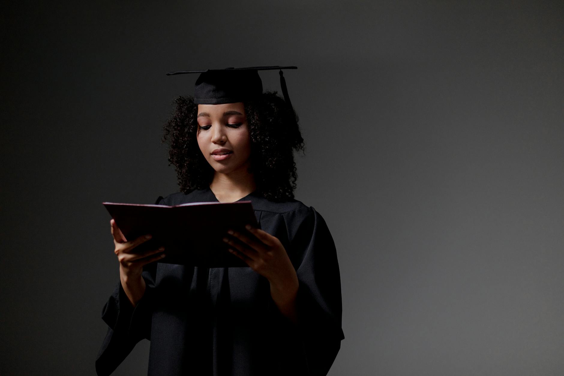 A graduate in cap and gown reads a book against a neutral background, symbolizing achievement and education. - post grad relationships