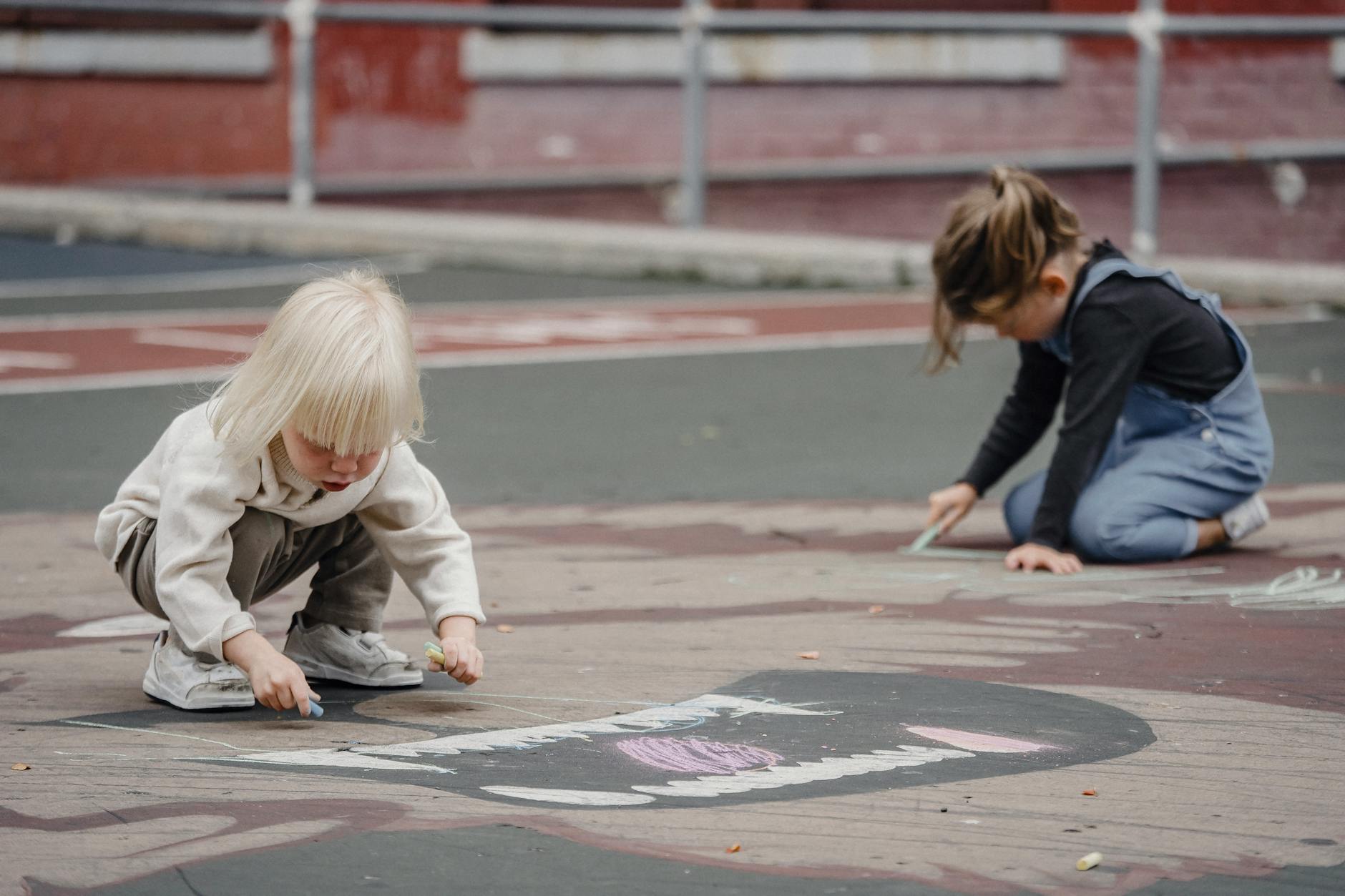 Side view of full body cute concentrated little girls drawing on asphalt ground with chalks - preschool boundaries outdoors