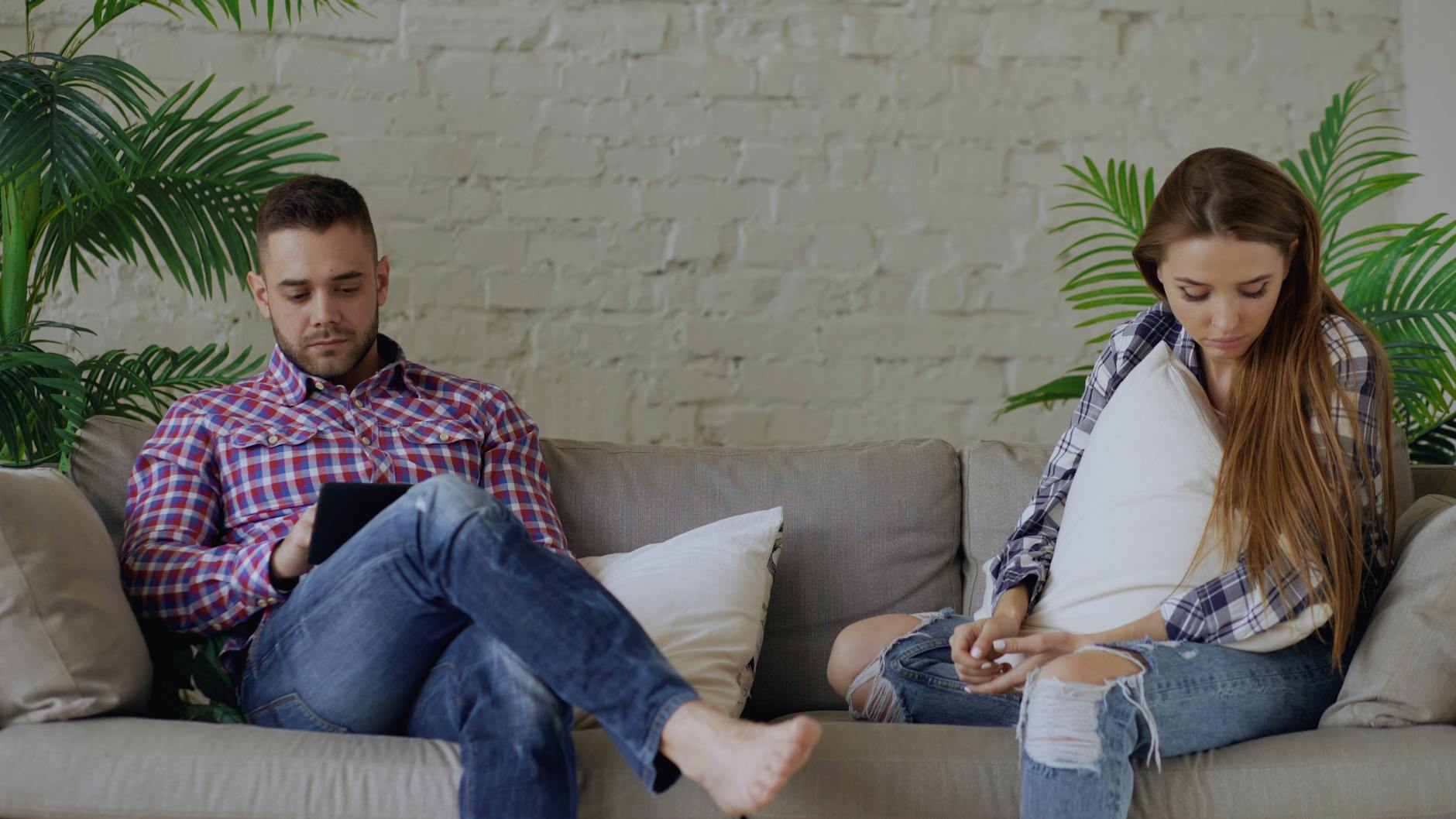 A couple sits apart on a couch, engrossed in their thoughts or devices in a casual living room setting. - relationship boredom therapy