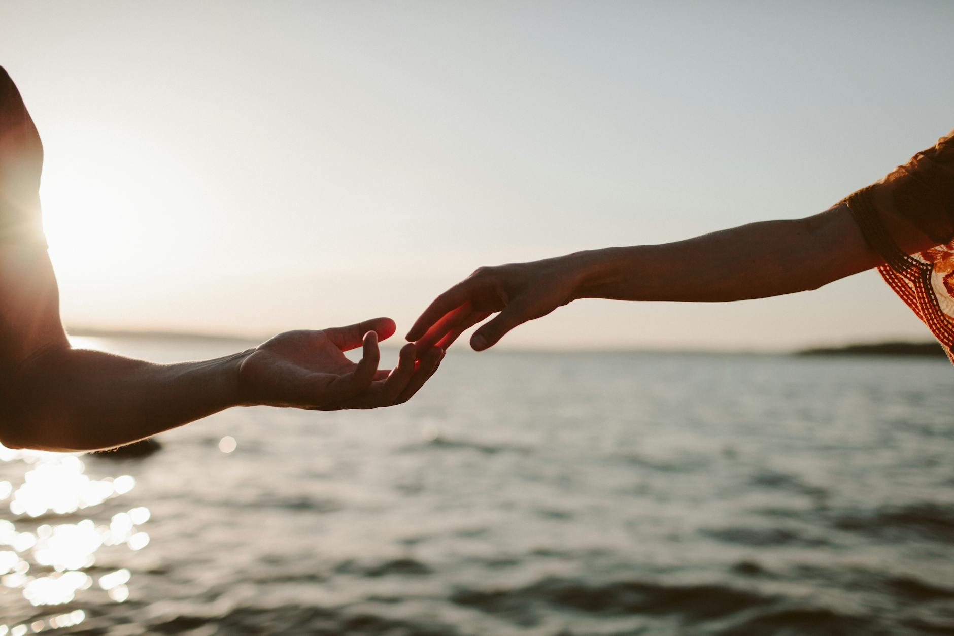 Silhouettes of hands reaching out at sunset over a tranquil sea, symbolizing connection and longing. - relationship check in