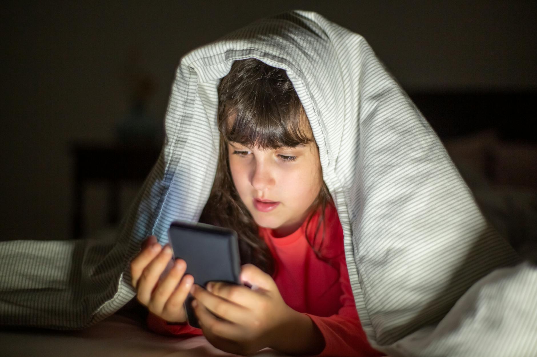 Young girl under blanket focused on a smartphone's glowing screen in a dim room. - screen time limits