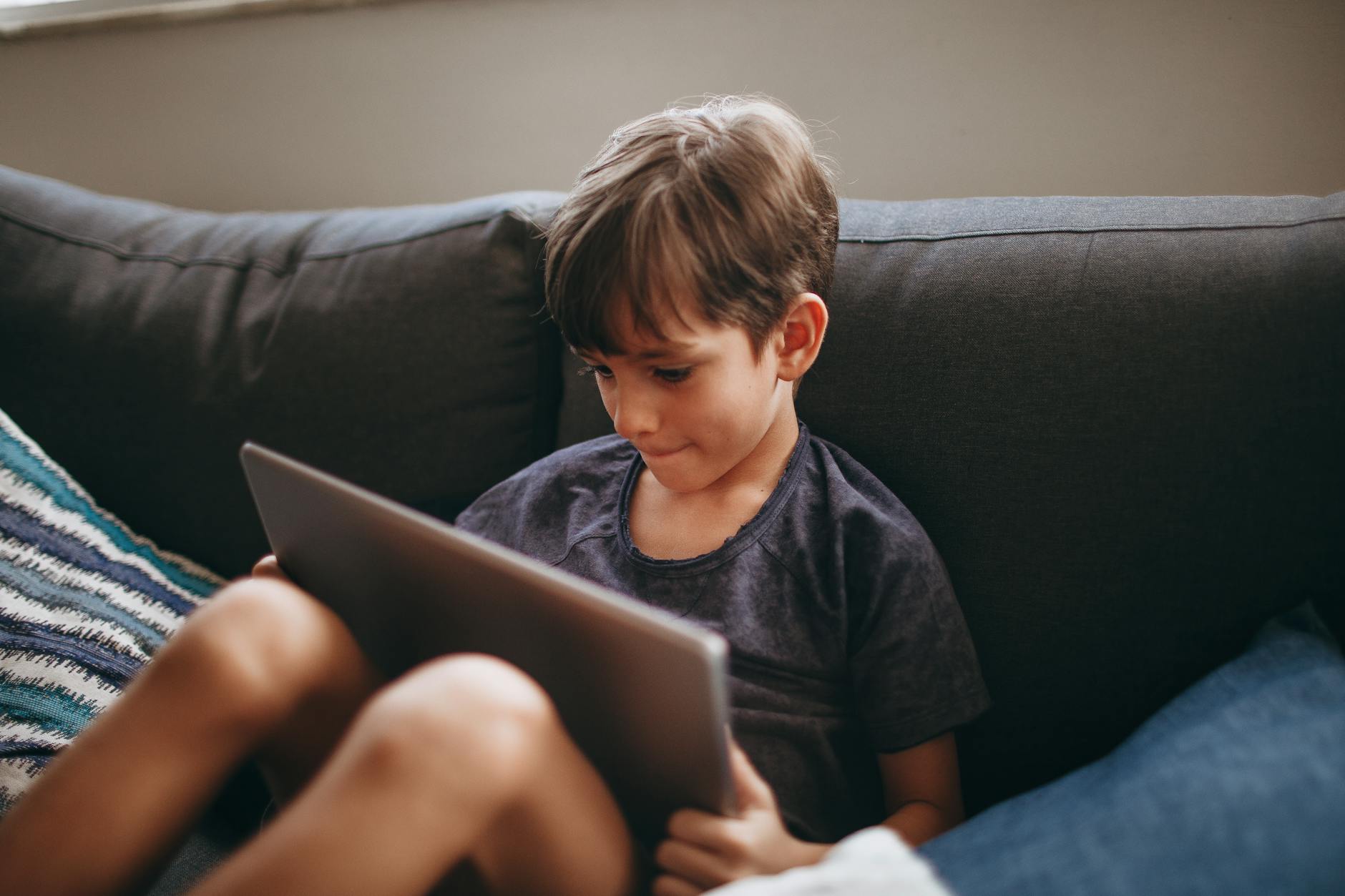A young boy attentively using a tablet while sitting on a comfortable sofa indoors. - screen time limits
