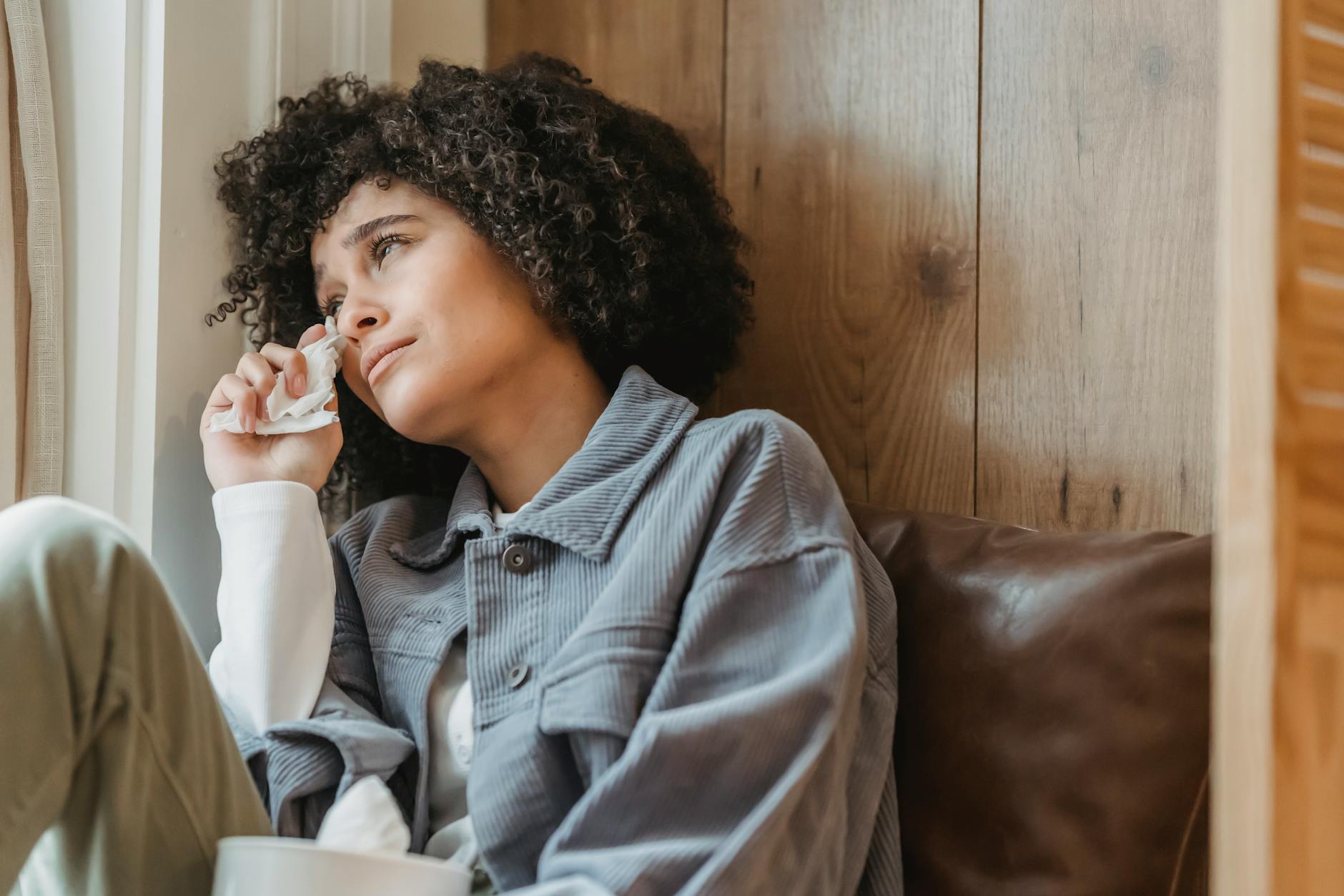 Desperate African American female with curly hair wiping face with tissue while crying from sadness on windowsill and looking away - seasonal depression treatment