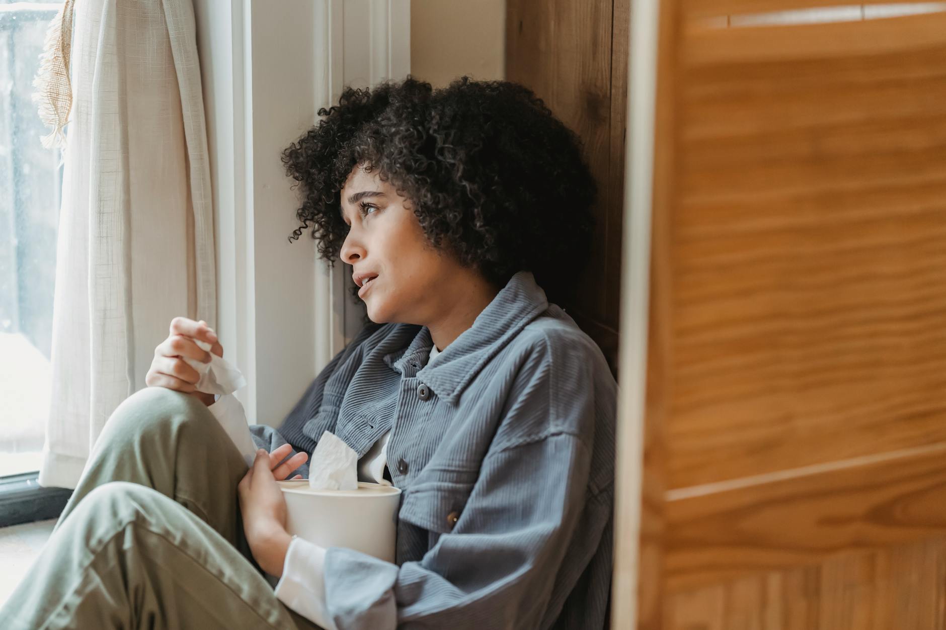 Side view of upset African American curly haired female crying on windowsill with box of tissues - seasonal depression treatment