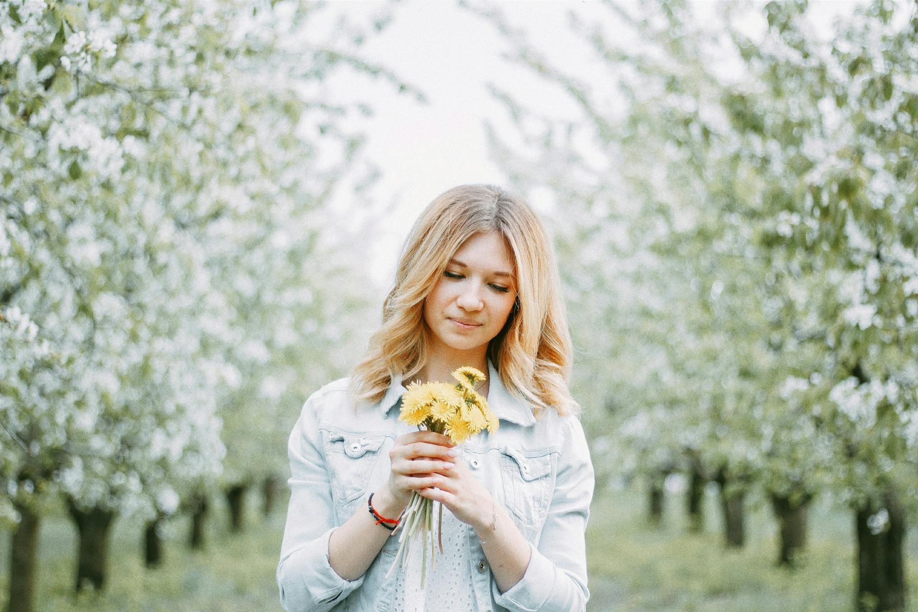 A young woman with blond hair holds dandelions in a blooming orchard, signifying springtime beauty. - spring fever anxiety