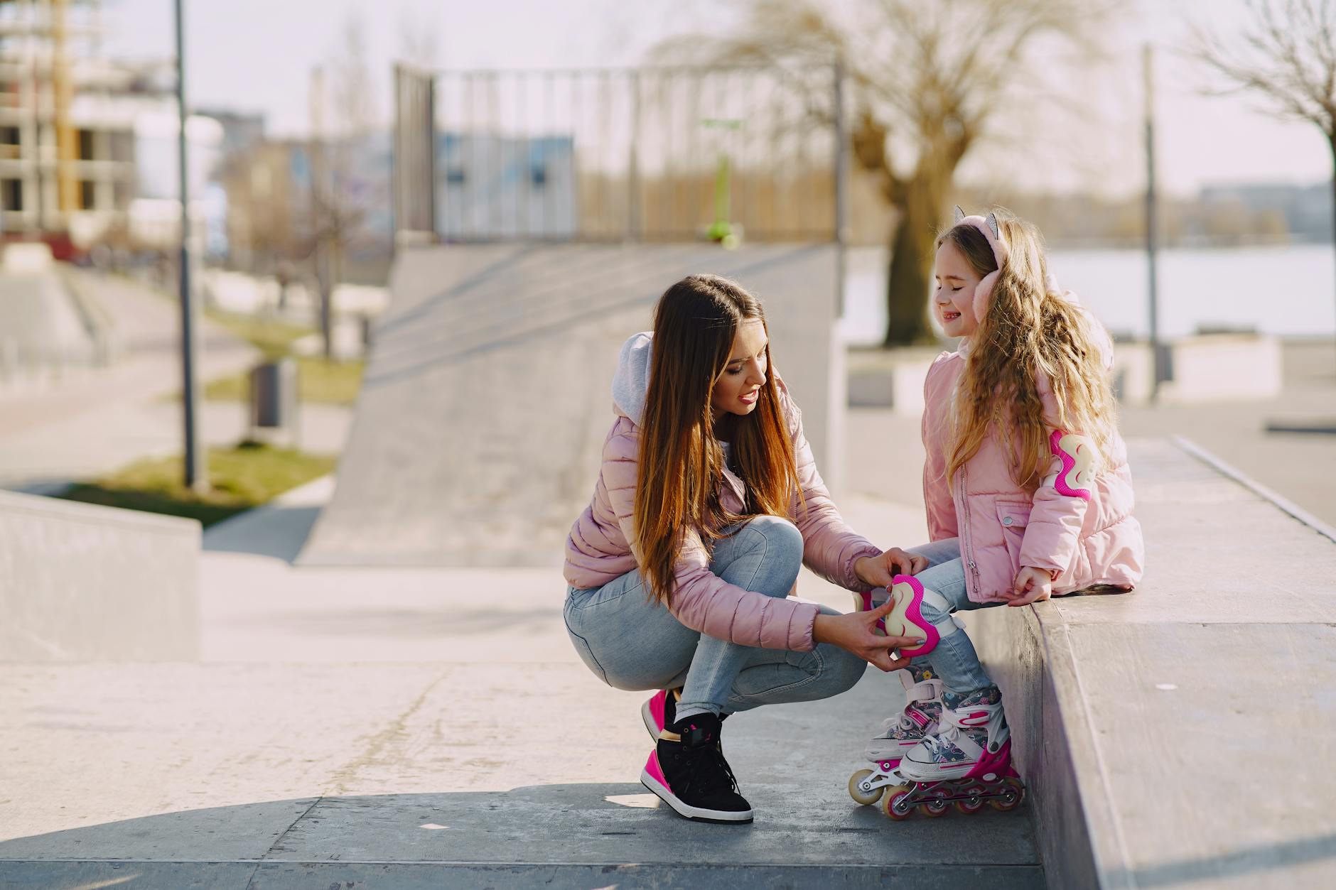 Cheerful young woman adjusting knee pads for little girl during active sport and game outside in city while spending time together - spring outdoor safety