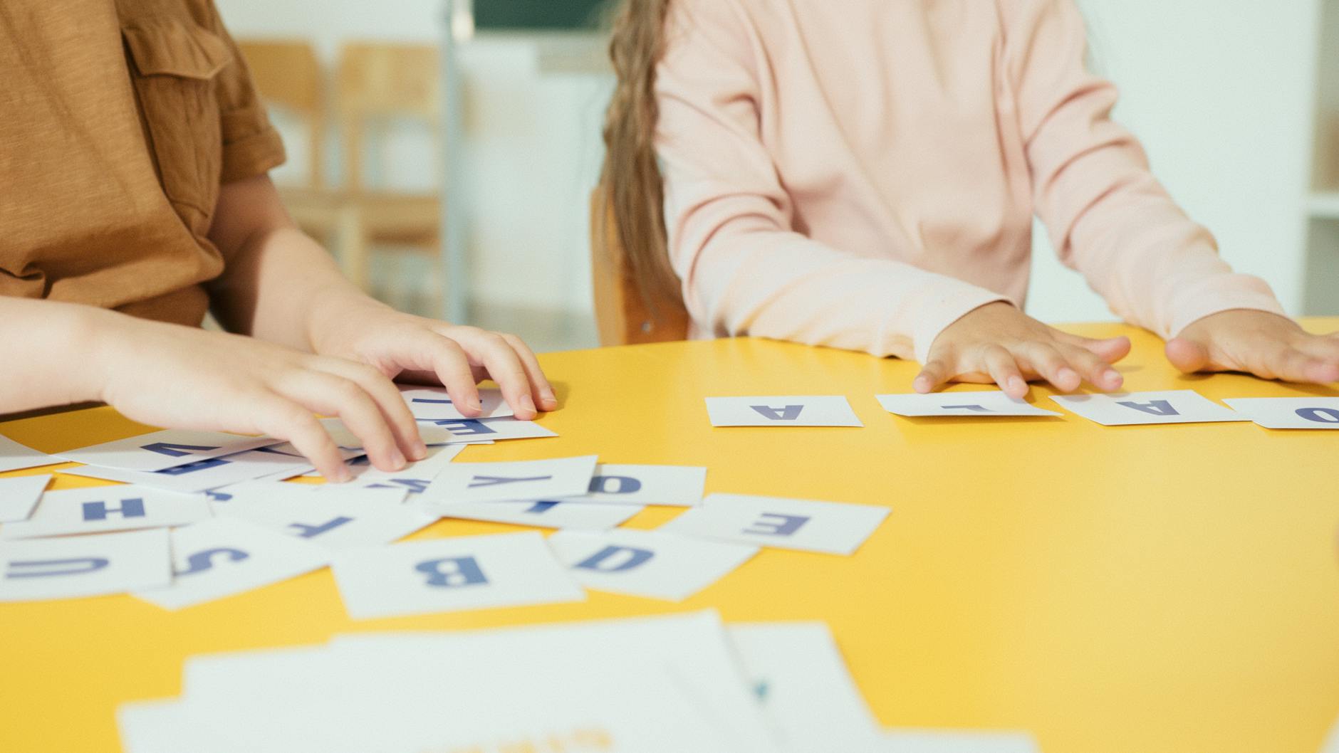 Children arranging alphabet cards on a yellow table in a classroom setting. - summer learning activities