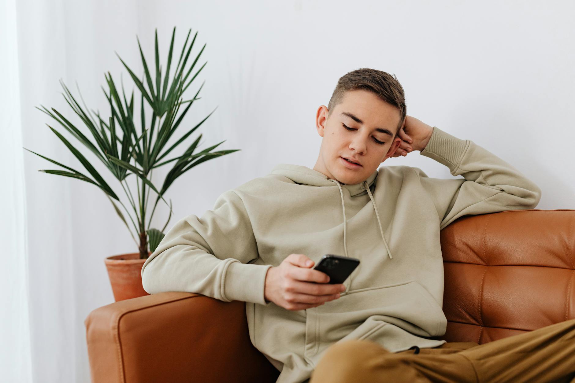 Teenage boy sitting indoors texting on a smartphone, wearing a hoodie. - teen social pressure