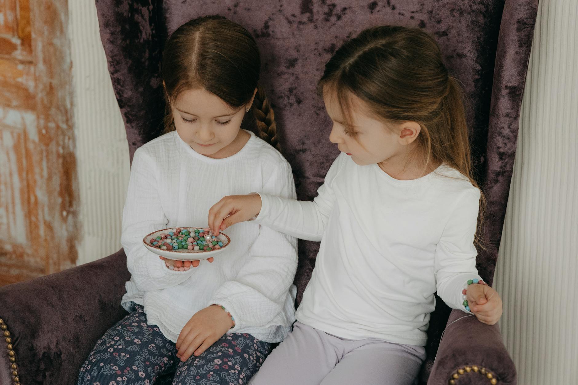 Two young girls sharing colorful beads while sitting on a purple armchair indoors. - toddler sharing