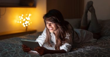 Beautiful woman lying in bed and holding tablet computer, chatting online using dating apps while relaxing at home late at night