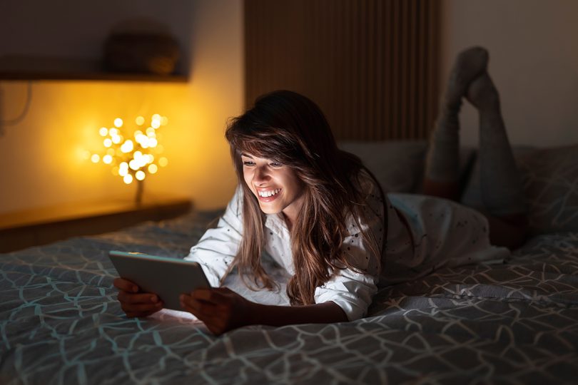 Beautiful woman lying in bed and holding tablet computer, chatting online using dating apps while relaxing at home late at night